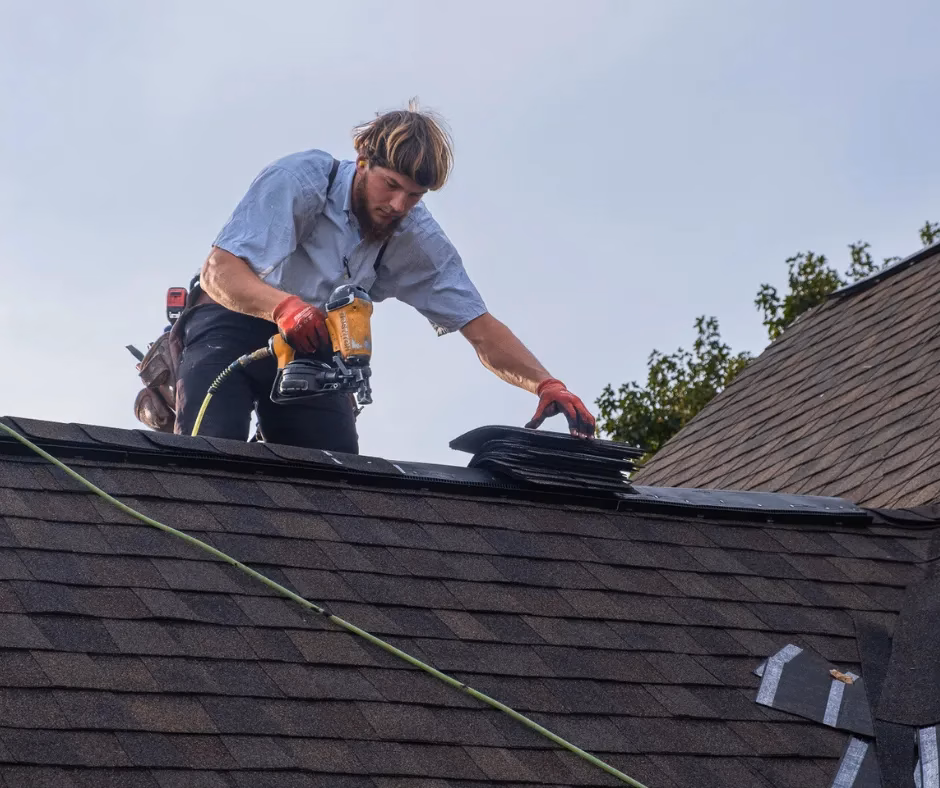 Man using a nail gun to install roofing shingles on a sloped roof, showcasing skilled labor and home improvement services.